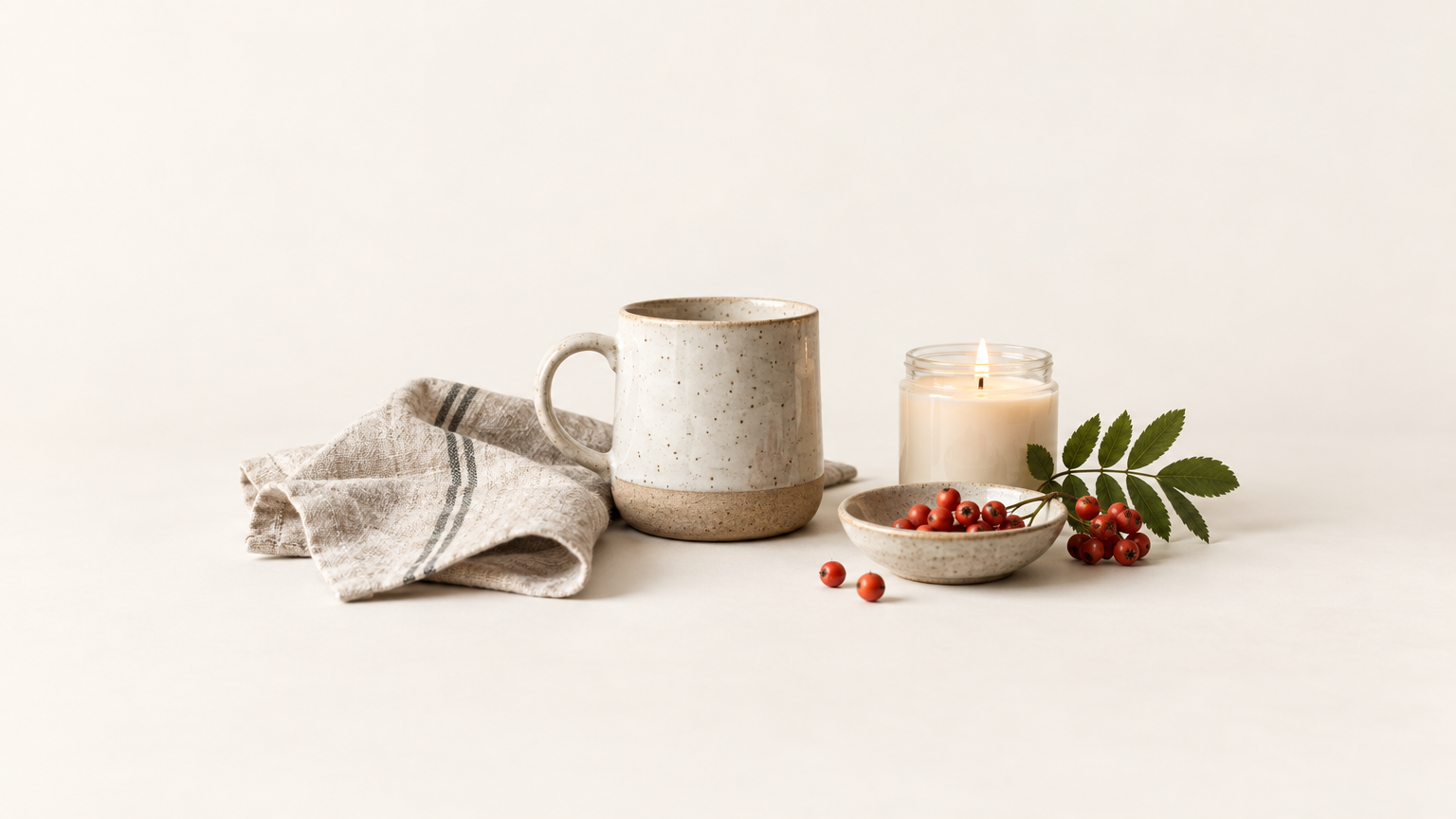 A table of maker goods: linen, a ceramic mug, a candle, and rowan berries