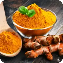 Bowl and spoon of turmeric powder next to turmeric roots and basil leaves.