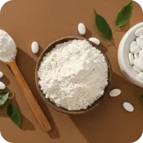 A bowl of white powder next to a spoon and white beans, with green leaves scattered around.