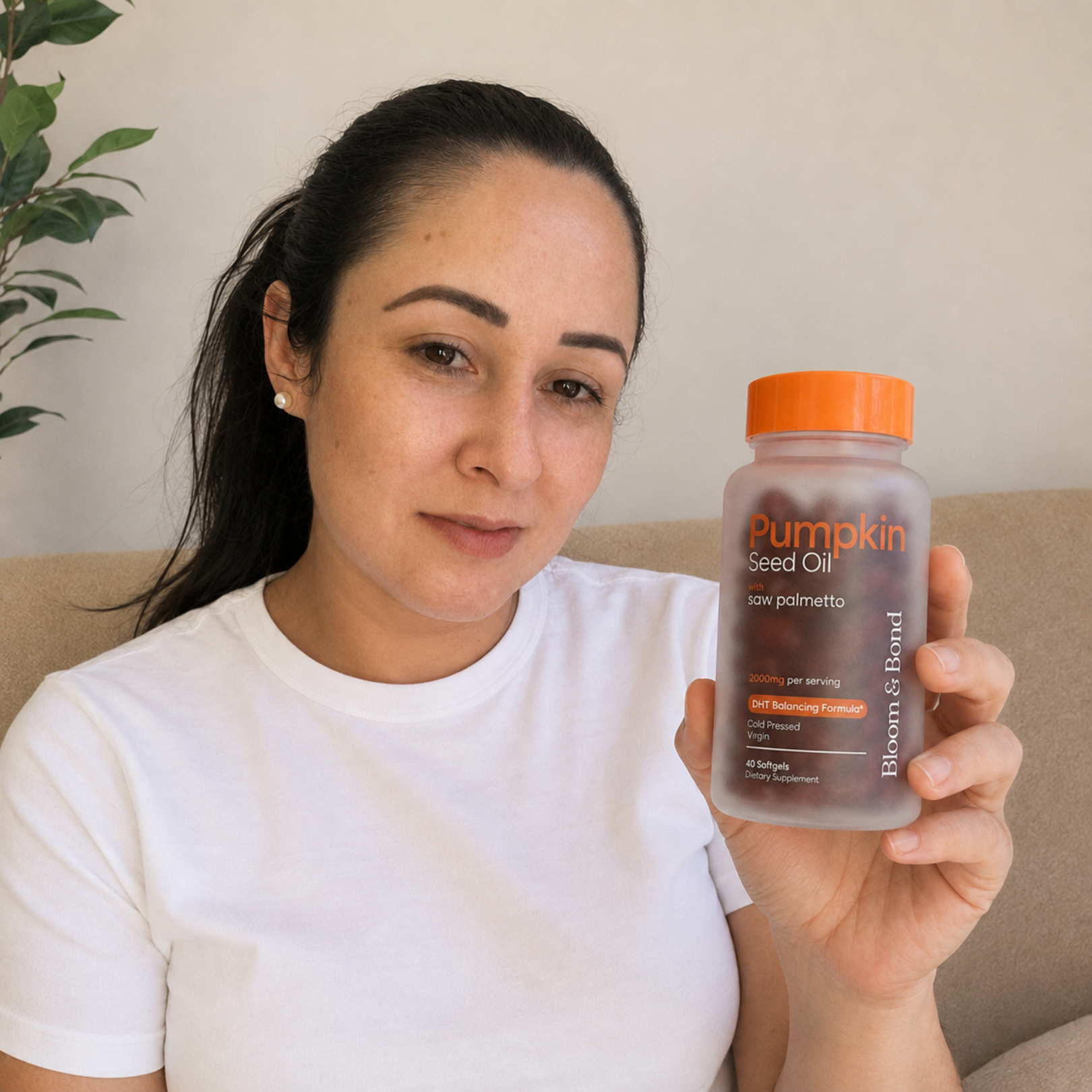 A woman in a white t-shirt sits on a couch holding up a bottle of Pumpkin Seed Oil supplements.