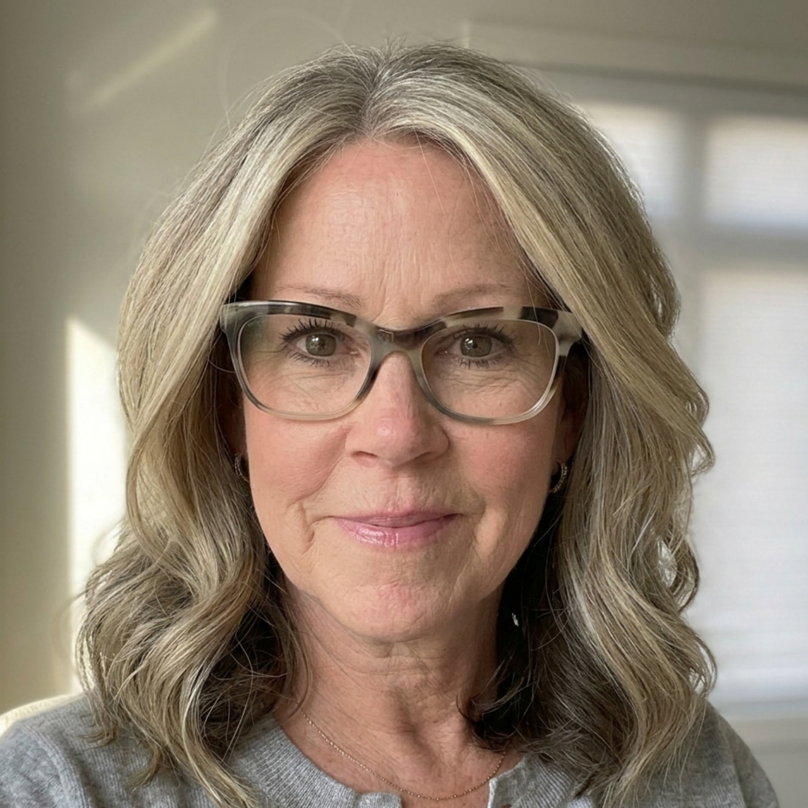 A headshot of a smiling woman with wavy, graying hair and patterned glasses, wearing a gray sweater.