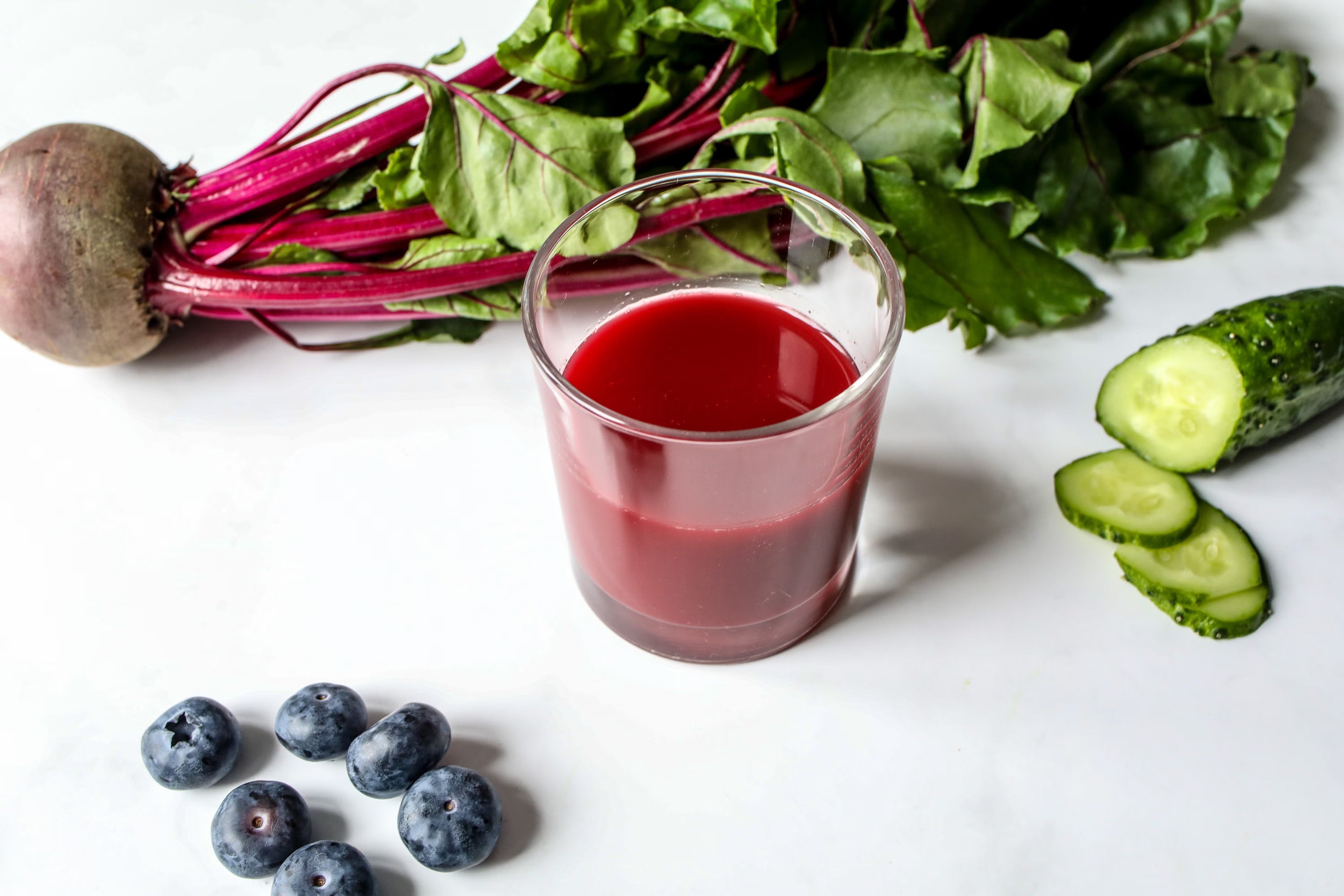 Cup of beetroot juice encompassed by fully intact betroot plant at top, cucumber to right, and blueberries to bottom left on white table surface.