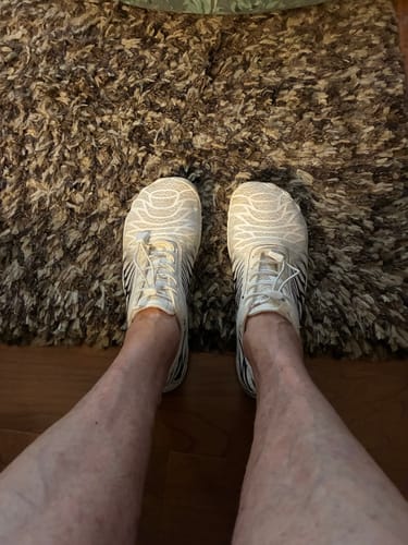 Reviewer looking down at their feet in white patterned Lorax Pro barefoot shoes while standing on a rug.