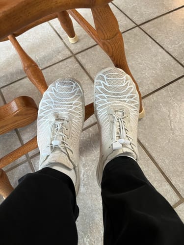 Reviewer's feet wearing beige Lorax Pro barefoot shoes with white lace-up toggles while sitting on a wooden chair.