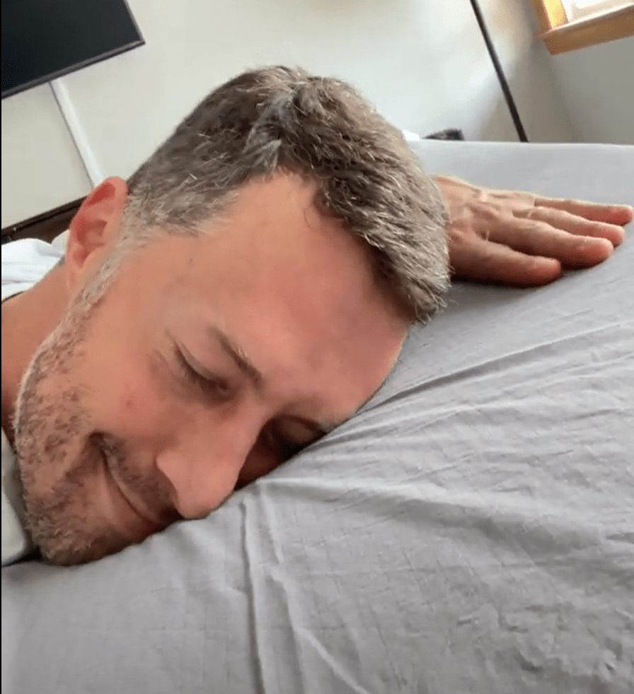 A man lies face-down on a bed, resting his head against the sheet.