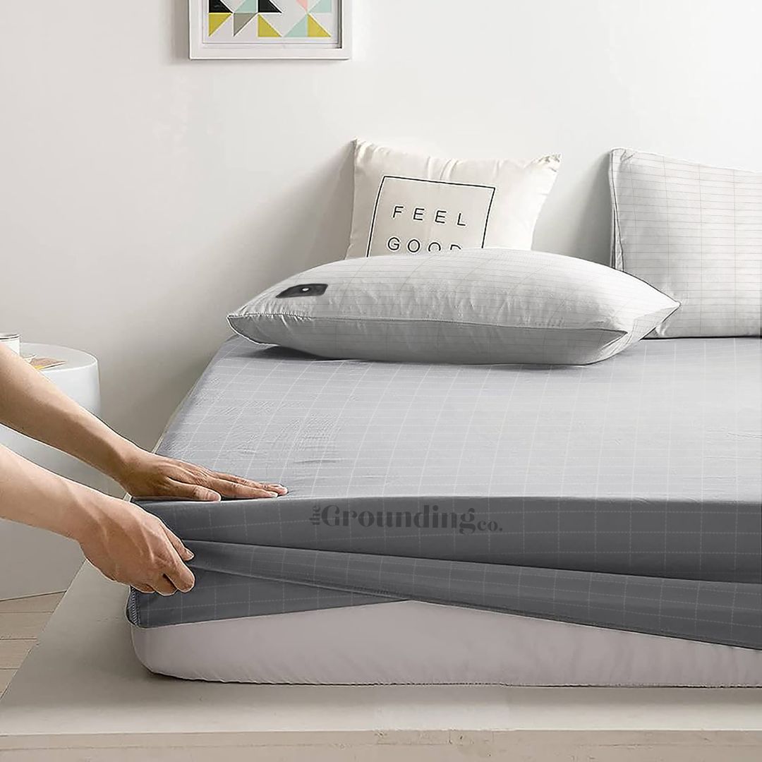 A neatly made bed with gray bedding and a 'FEEL GOOD' cushion, and a hand pulling the fitted sheet corner.